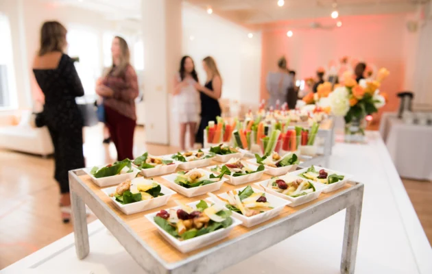 Elegant corporate lunch event with beautifully plated appetizers, fresh salads, and colorful vegetable shooters on a stylish buffet table, with guests mingling in the background.