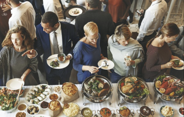 A diverse group of professionally dressed people selecting food from a buffet table with gourmet dishes at a corporate event.