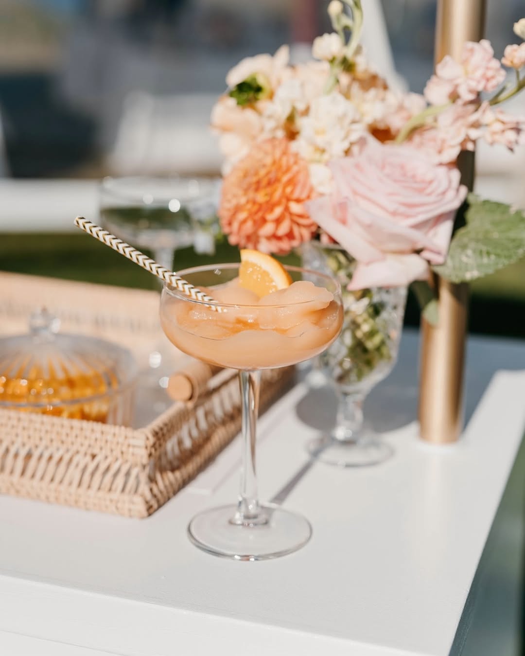 Peach-colored signature cocktail garnished with citrus and served beside soft pastel floral arrangements at an outdoor summer celebration.